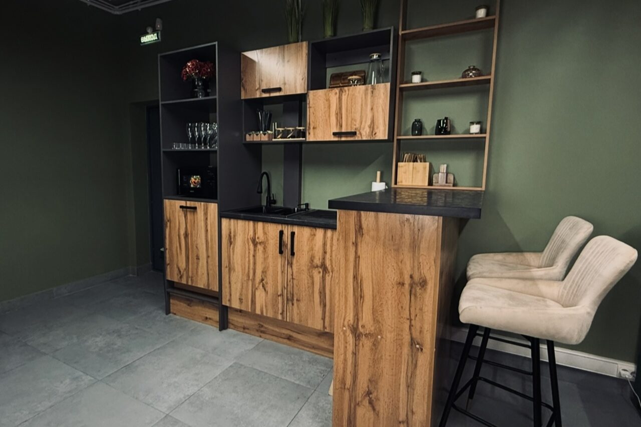 Арендовать Лофт Интра. Зал Фрей. Современная компактная кухня с барной стойкой. Деревянные шкафы, темная столешница, встроенная раковина. Рядом барные стулья с мягкой обивкой. Пол серый плиточный, стены зелёные. Открытые полки с декором и посудой. Верхнее освещение.