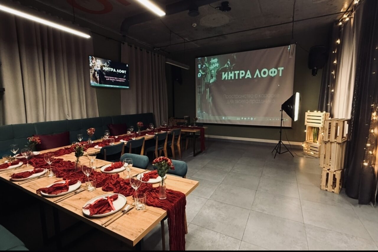 Арендовать Лофт Интра. Зал Фрей. Лофт-пространство с высоким потолком и серым полом. Слева длинный деревянный стол с сервировкой для ужина, красные декорации. Справа проекционный экран и стул. Светильники на потолке. На стене вывески на русском языке.
