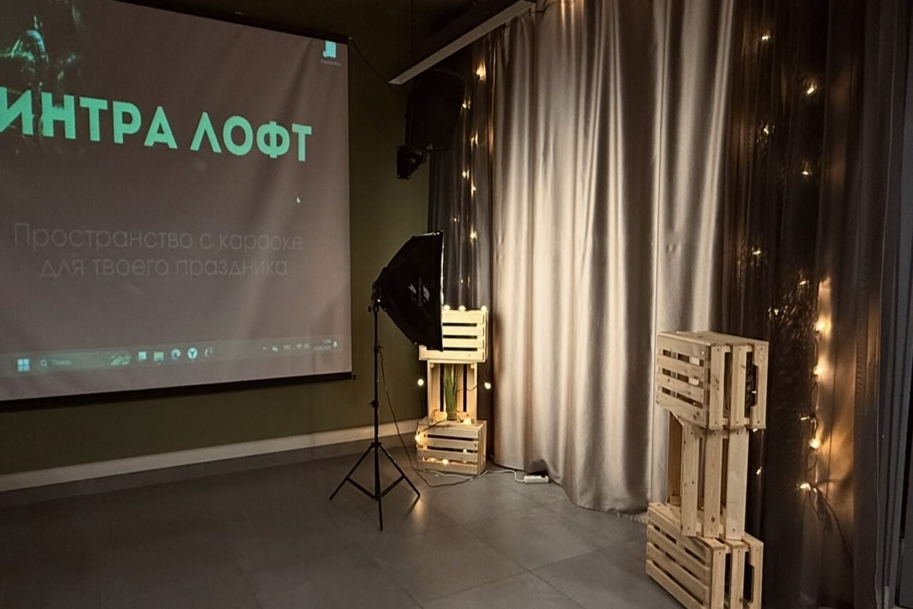 Арендовать Лофт Интра. Зал Фрей. Лофт-пространство с проектором на зеленой стене, серыми шторами, стулом из деревянных паллет и мягким освещением. Стены и пол темного цвета, светящиеся гирлянды вдоль штор. Текст на экране: "ИНТРА ЛОФТ Пространство".