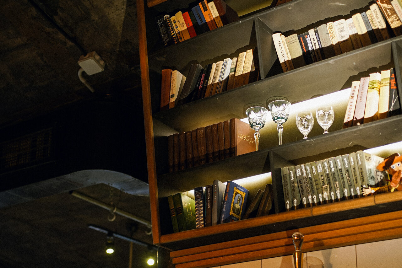 Арендовать Гастробар Дневник. Настенная полка с книгами и бокалами, освещённая тёплым светом. Верхняя часть полки заполнена книгами в разноцветных обложках. На полке также стоят бокалы для вина. Потолок с трубами и софитами.