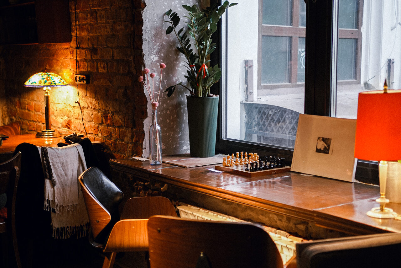 Арендовать Гастробар Дневник. Кофейня с кирпичной стеной, деревянной стойкой у окна, настольными лампами с цветными и красными абажурами. На стойке шахматная доска, декор из растений и сувенирных фигурок. За столом несколько деревянных стульев. Естественное освещение из окна.