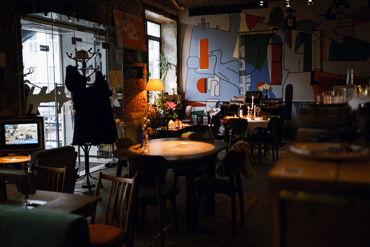 Арендовать Гастробар Дневник. Кафе с круглыми деревянными столами и стульями, настенные росписи, лампа на столе, вешалка с одеждой, за окном видна улица, приглушенное освещение, на столах свечи и бокалы, бетонные стены, тёплые цвета.