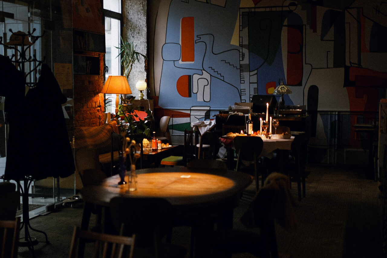 Арендовать Гастробар Дневник. Интерьер кафе с мебелью из дерева. Круглые и квадратные столы, стулья. На стенах абстрактные рисунки. Освещение мягкое, с настольными лампами и свечами. Пол темный, ковровое покрытие. Есть вешалки для одежды и комнатные растения.