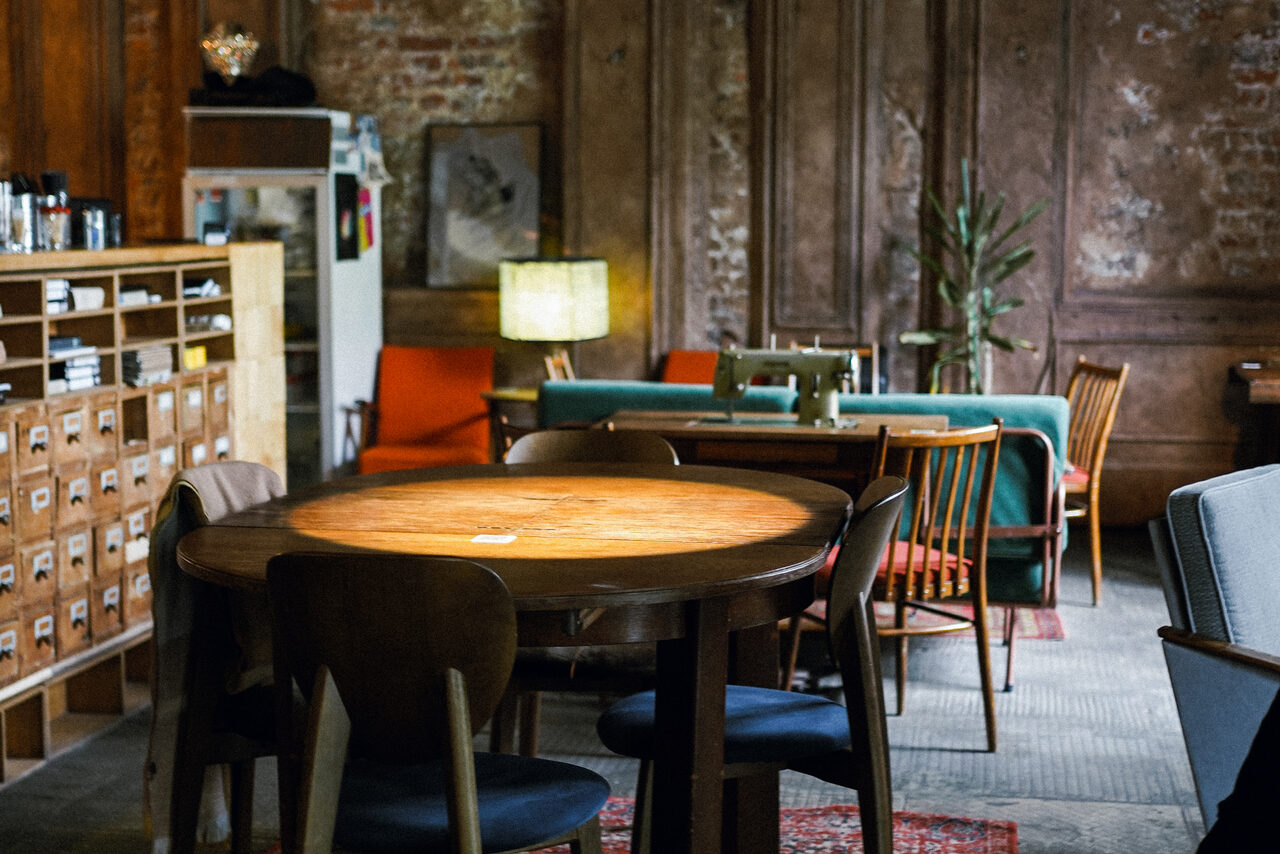 Арендовать Гастробар Дневник. Теплое, уютное пространство с круглым деревянным столом и стульями, вмещающее диваны и кресла. Стена с кирпичной отделкой, книжный стеллаж. Естественное освещение из лампы, ковры на полу. Оформлено в ретро-стиле с растениями.