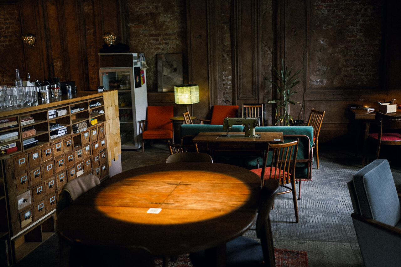 Арендовать Гастробар Дневник. Кофейня с деревянными стенами и кирпичной кладкой, лампы, деревянные столы и стулья, мягкие кресла, стойка с посудой и полками, шкафчики с ящиками, лампа с абажуром, рассеянное освещение, ковровое покрытие, зелёное растение.