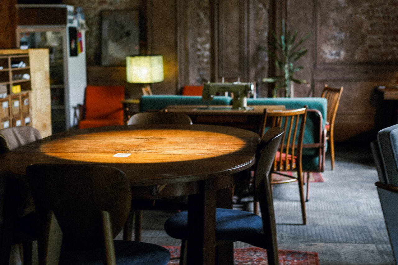 Арендовать Гастробар Дневник. Просторное помещение с круглым деревянным столом и стульями, швейная машина на другом столе, диван и кресла. Освещение от лампы с абажуром. Декор из кирпичных и деревянных панелей. Цвета: коричневый, оранжевый, бирюзовый.