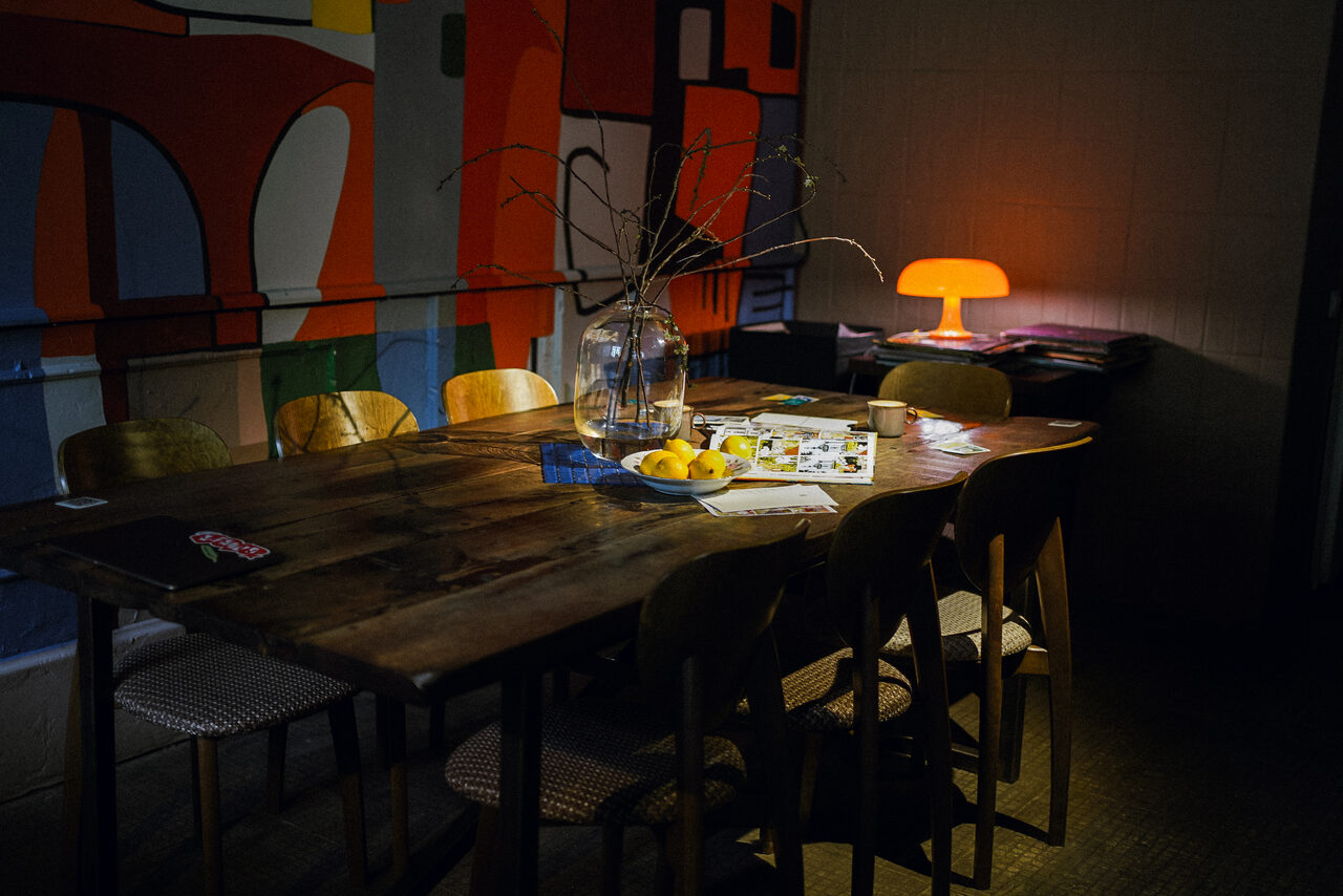 Арендовать Гастробар Дневник. Пространство с деревянным столом и восьмью стульями, стеклянная ваза с ветками на столе, рядом журналы и лимоны. На стене яркий абстрактный рисунок. В углу стол с оранжевой лампой. Освещение мягкое, создаёт уютную атмосферу.