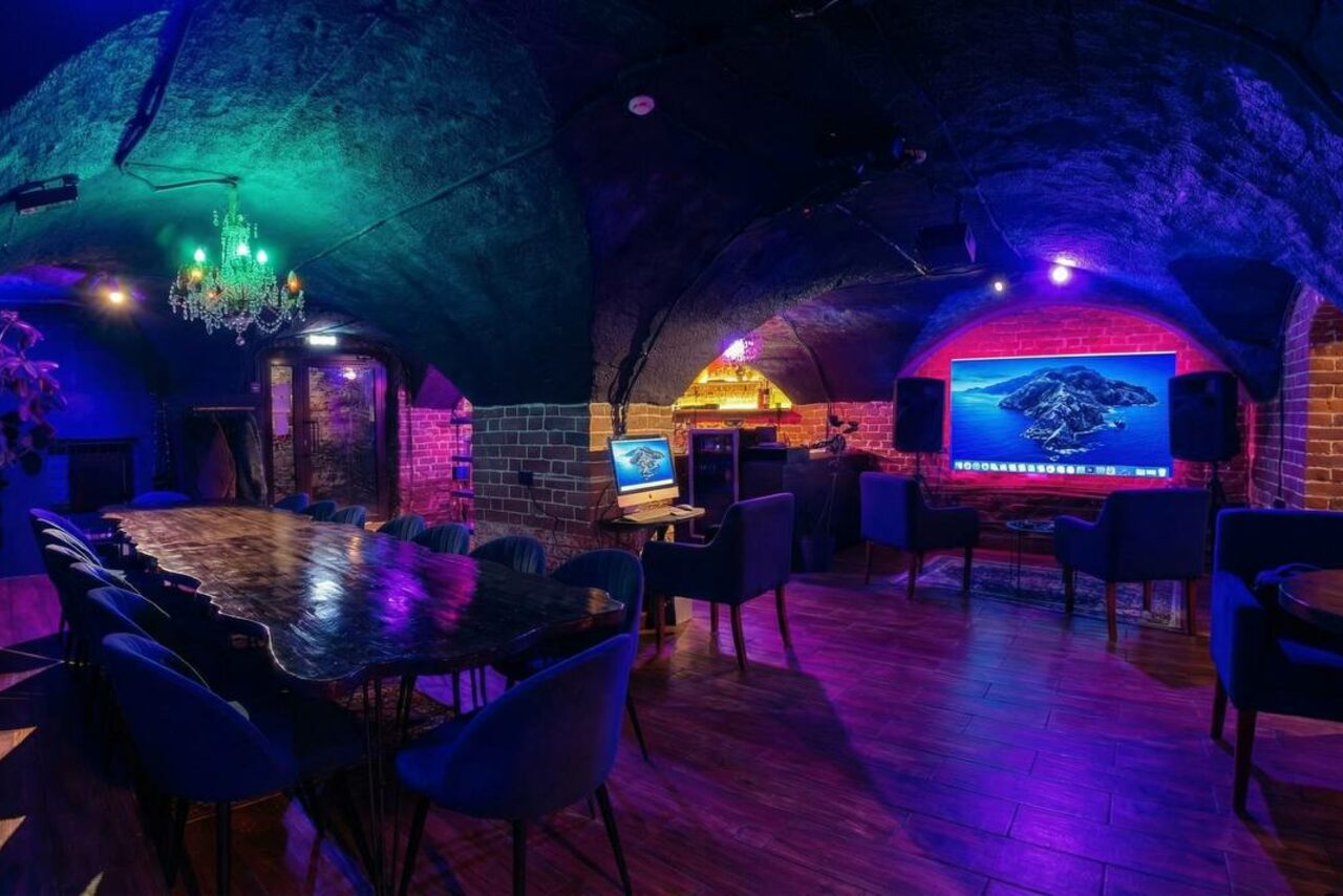 Арендовать GB Rooms(ГБ Румс). Лофт №1. Подвальное помещение с кирпичными стенами и арочными сводами. Длинный деревянный стол с мягкими стульями. На стене большой экран и компьютерный монитор. Фиолетовое и зеленое освещение, подвесная люстра. Деревянный пол, динамики по бокам экрана.