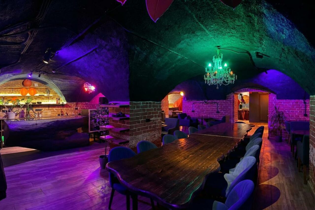 Арендовать GB Rooms(ГБ Румс). Лофт №1. Подвальное помещение с кирпичными стенами и арочным потолком. Длинный деревянный стол и мягкие стулья. Барная стойка в глубине. Цветное освещение: зелёные и фиолетовые оттенки. Люстра в центре. На полу деревянное покрытие.