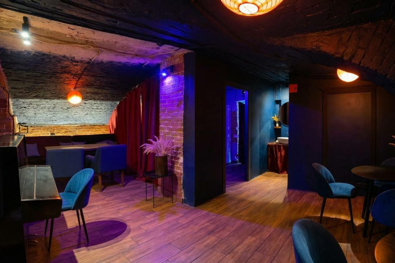 Арендовать GB Rooms(ГБ Румс). Лофт №1. Интерьер помещения: кирпичные стены, деревянный пол, черный и синий цвет стен, закрыто красной занавесью. Мебель: синие кресла, столы, пианино. Освещение: теплые светильники, потолочные лампы. Декор: вазы с растениями.