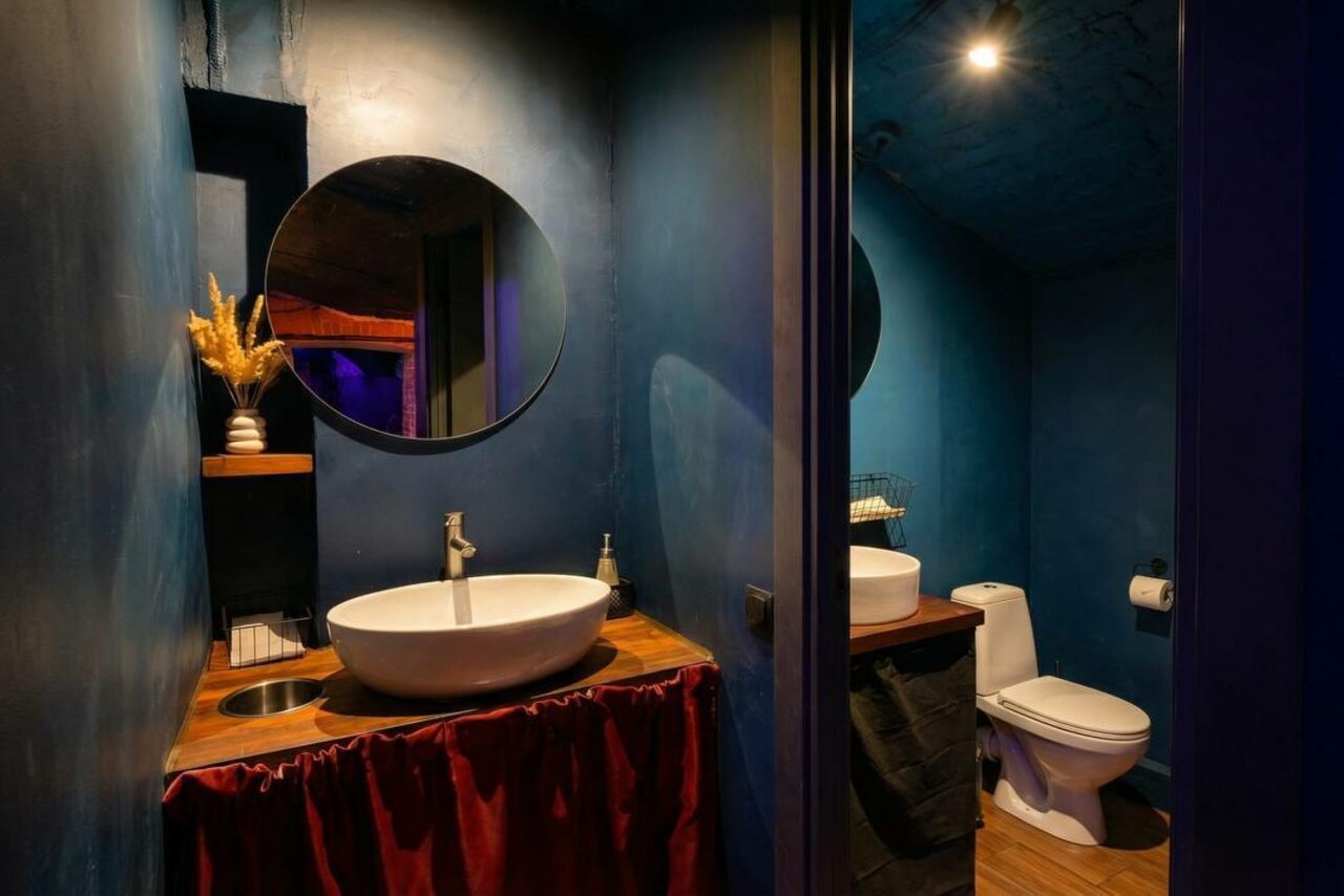 Арендовать GB Rooms(ГБ Румс). Лофт №1. Туалетная комната с темно-синими стенами, раковиной на деревянной тумбе с красной занавеской, круглым зеркалом, светильником, туалетом и декоративной вазой на полке. Освещение мягкое, материалы деревянные и текстильные.