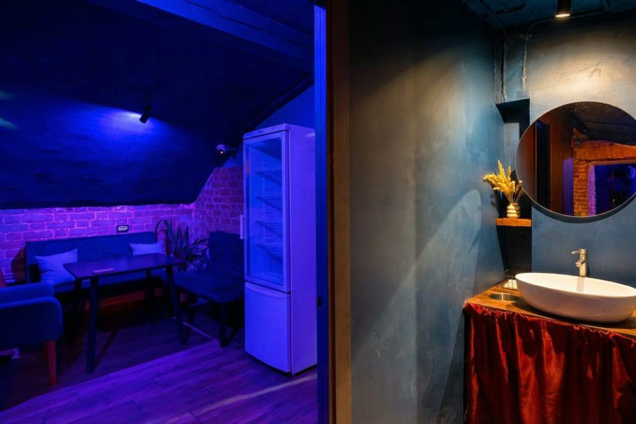 Арендовать GB Rooms(ГБ Румс). Лофт №1. Помещение с зоной отдыха и санузлом. В зоне отдыха диван, стол, стул, холодильник, кирпичная и синяя стена. В санузле круглое зеркало, раковина, лампы, синяя стена, красная тканевая юбка под раковиной. Залито синим освещением.