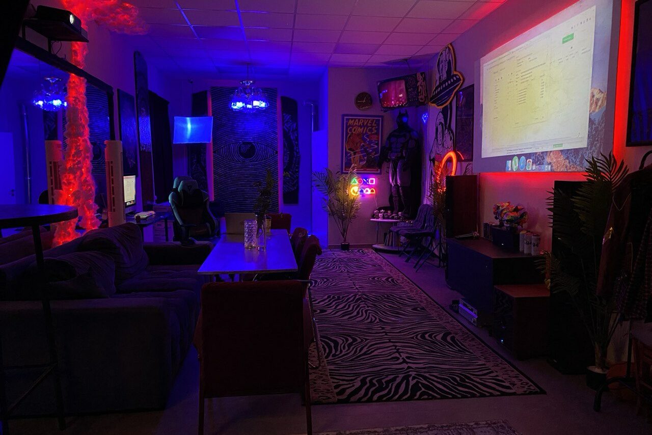 Арендовать GB Rooms(ГБ Румс). Лофт №2. Пространство с открытой планировкой, включающее зону отдыха с диваном и стульями, обеденный стол, проектор на стене, декоративный ковер с зебровым узором. Освещение разноцветное — красное и синее. Интерьер украшен постерами и растениями.