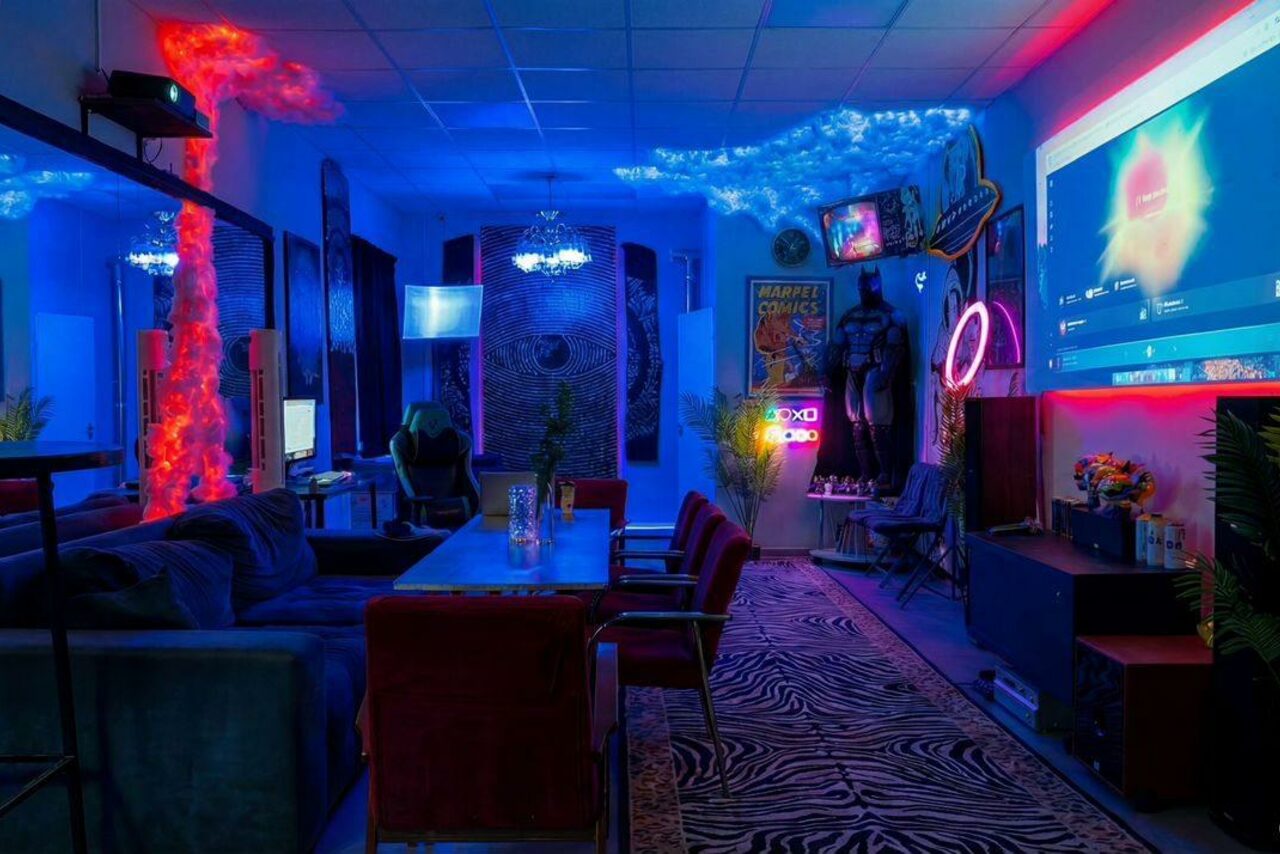 Арендовать GB Rooms(ГБ Румс). Лофт №2. Современная комната с диванами и креслами, столом, проектором на стене, разноцветной неоновой подсветкой, зеркалами, плакатами на стенах, растениями, декоративным освещением, акустической панелью и ковром с зебровым узором.