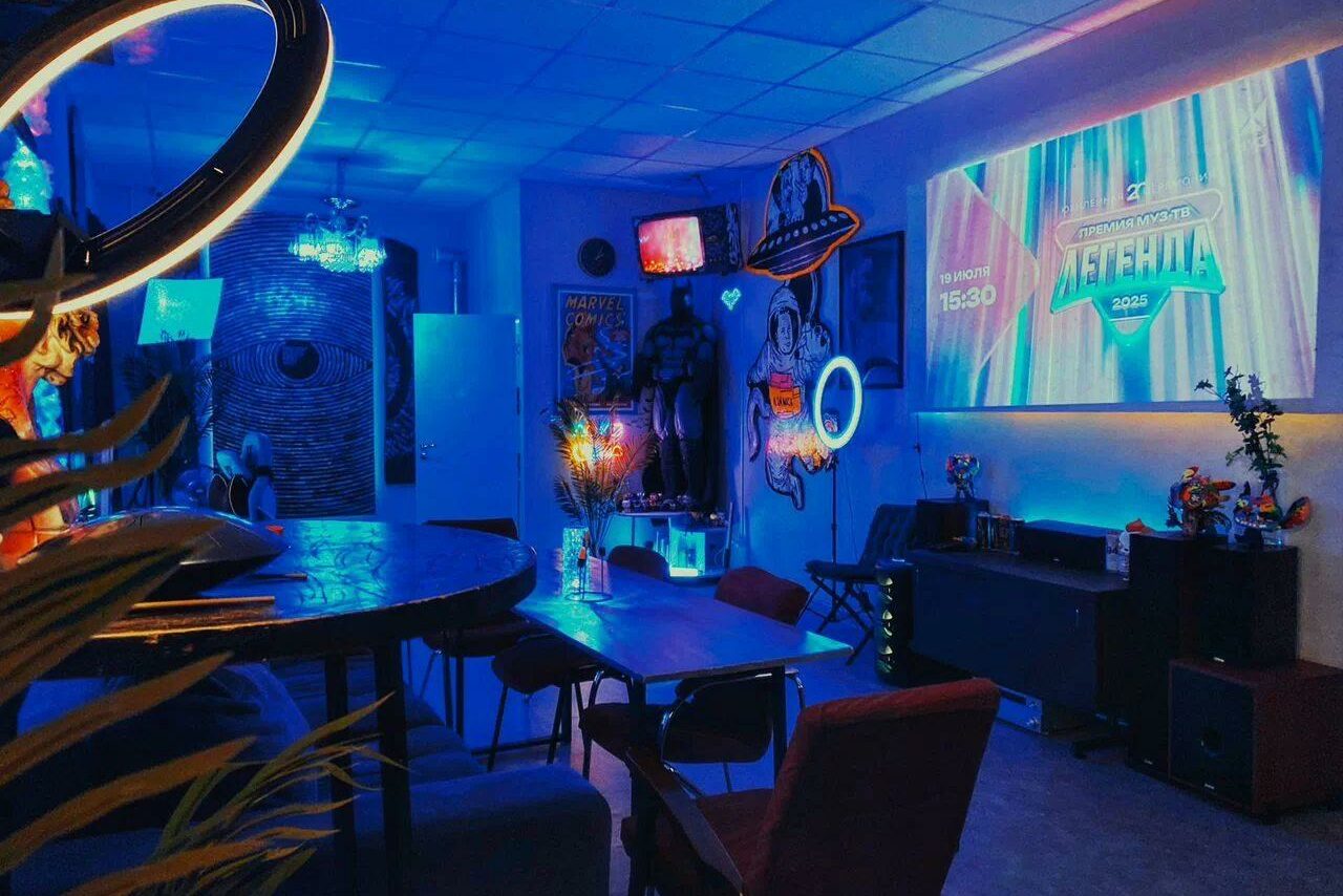 Арендовать GB Rooms(ГБ Румс). Лофт №2. Креативное пространство с неоновым освещением. В центре прямоугольный стол с стульями, рядом круглый стол со стульями. Неоновые знаки, проекция на стене. Декор в стиле комиксов, мягкая мебель, цветы в вазе. Игровая консоль на тумбе.