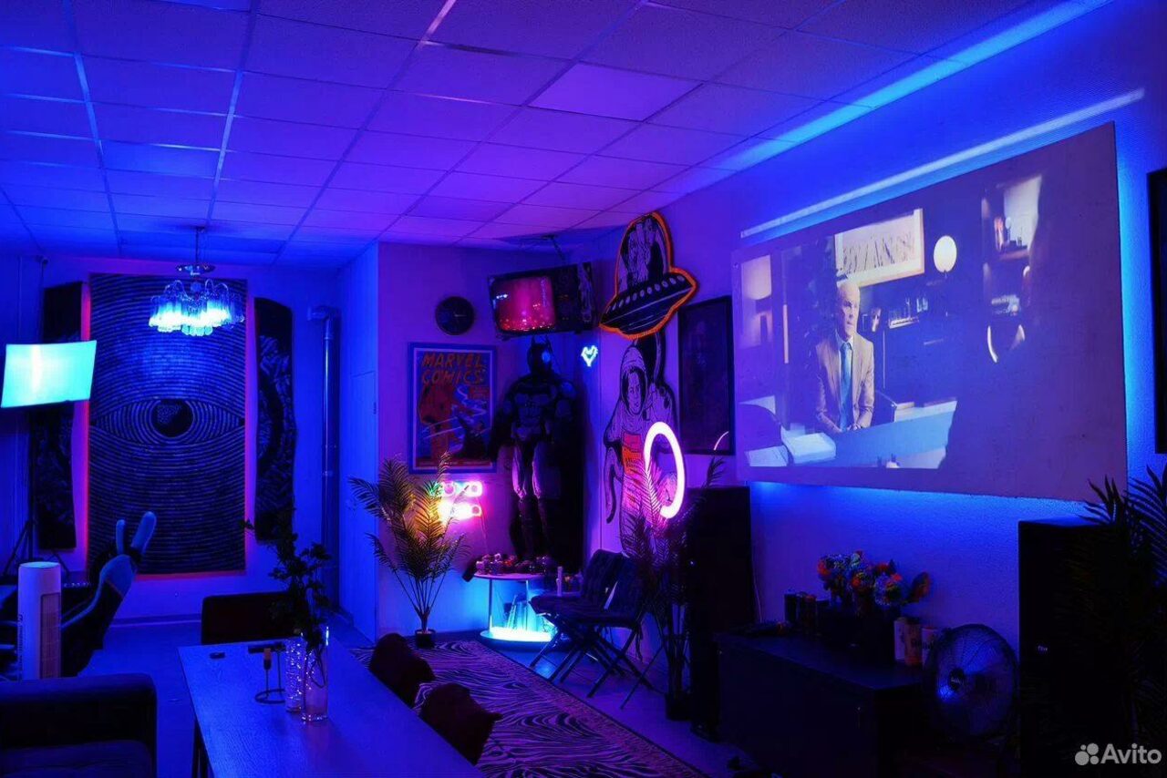 Арендовать GB Rooms(ГБ Румс). Лофт №2. Комната с синим и фиолетовым неоновым освещением, диванами вдоль стен, столом в центре. На стене проектируется изображение. Присутствуют декорации, неоновые вывески и растения. Потолок с плиткой, пол с ковром.
