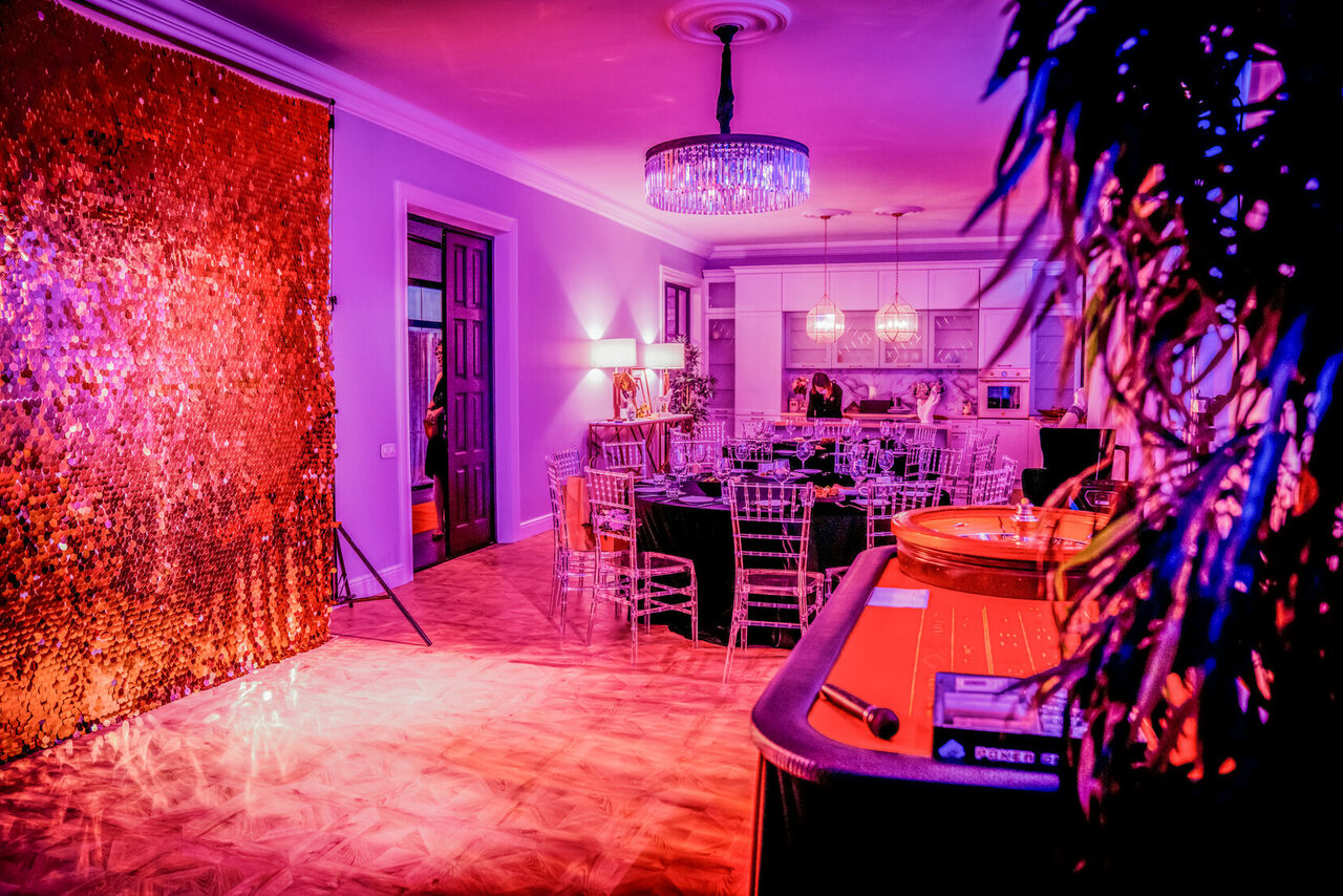 Арендовать Лофт Космос. Зал Президент. Пространство с зоной для чаепития и декором для мероприятий. Круглый стол с прозрачными стульями, кухня с барной стойкой, свисающие светильники. Стены украшены занавесом с блёстками. Подсветка фиолетового и оранжевого тонов.