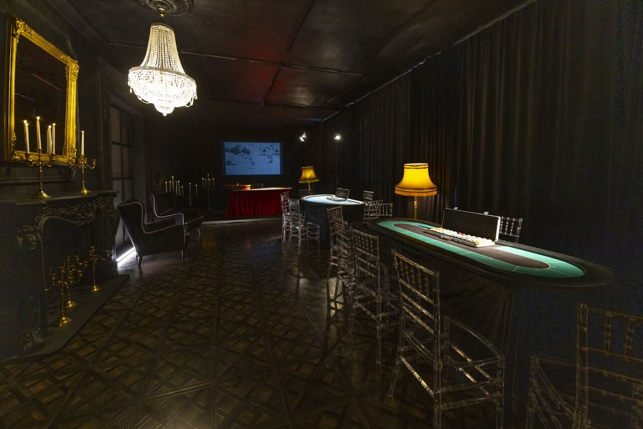 Арендовать Лофт Космос. Зал Президент. Игровое пространство с покерными столами, прозрачными стульями и классическими диванами. На стене проектор с изображением. Темные шторы, люстра и настольные лампы обеспечивают освещение. Паркетный пол, декоративные каминные элементы, зеркало и подсвечники.