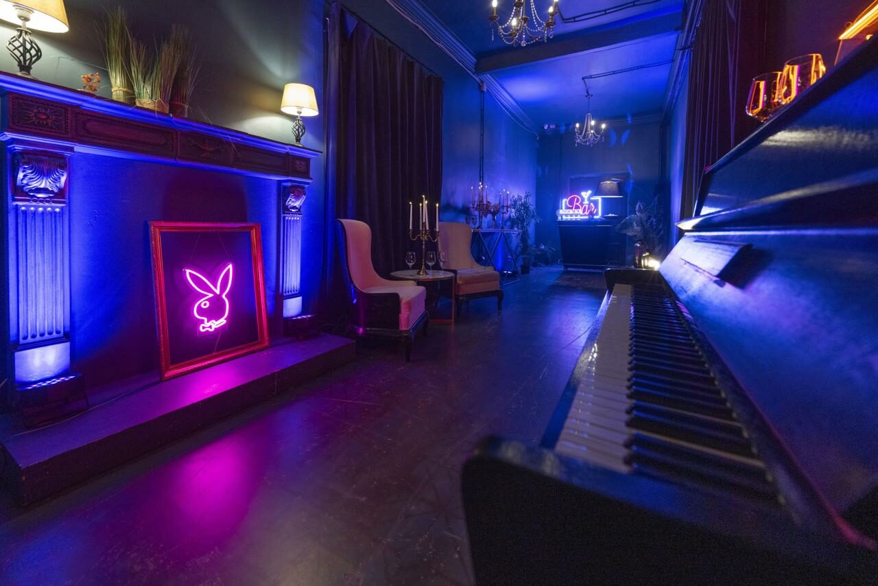Арендовать Лофт Космос. Зал Президент. Стильное пространство с пианино и мягкими креслами, украшенное неоновыми знаками. Темные стены, шторы и деревянный пол. Встроенные светильники и настенные бра создают атмосферное освещение. Декор в виде подсвечников и зелени на камине.