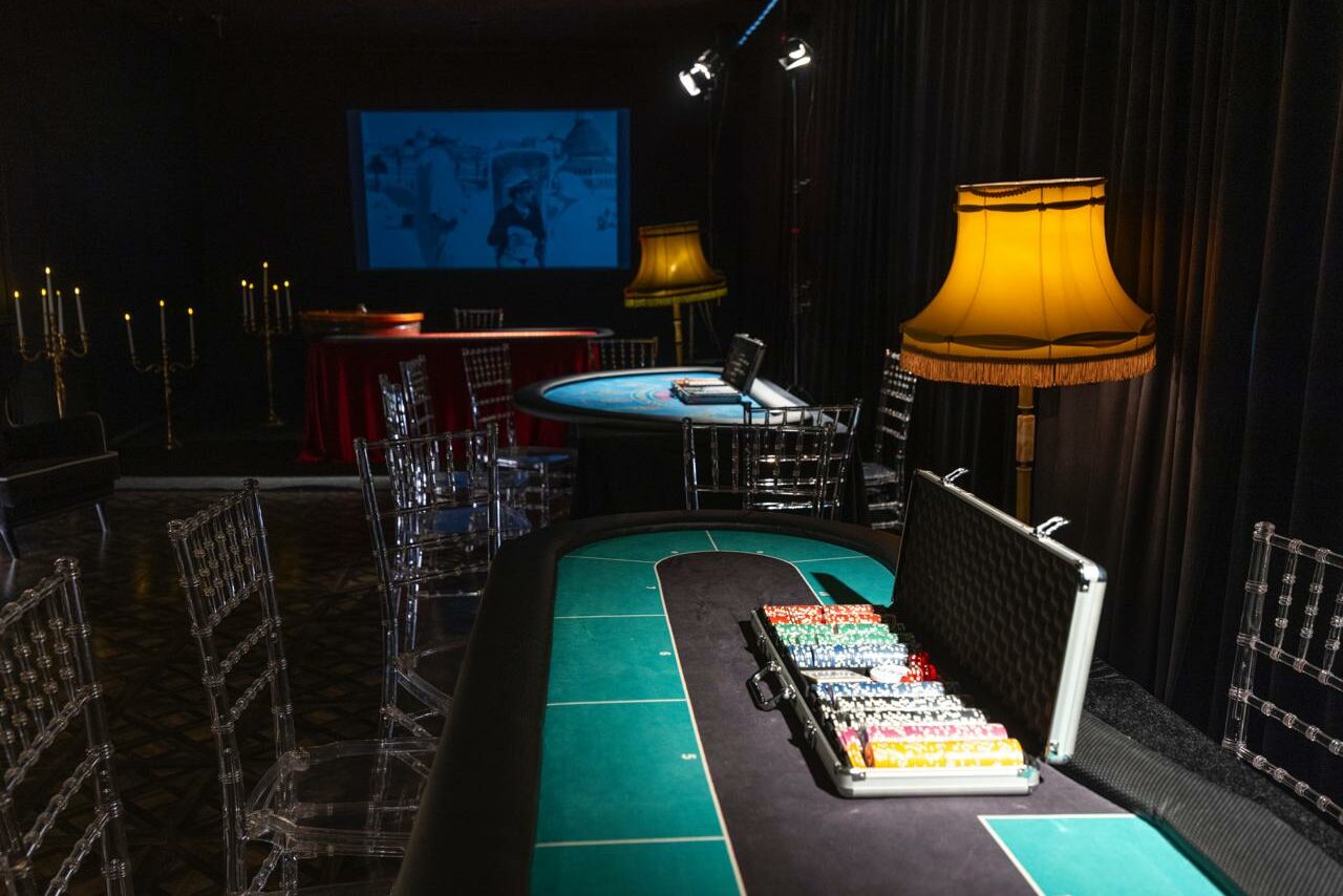 Арендовать Лофт Космос. Зал Президент. Комната с игорными столами, прозрачными стульями и черными занавесками. Настольные лампы с желтыми абажурами, кейс с фишками, проектор с экраном на стене, канделябры. Тёмное освещение, полы с узором.