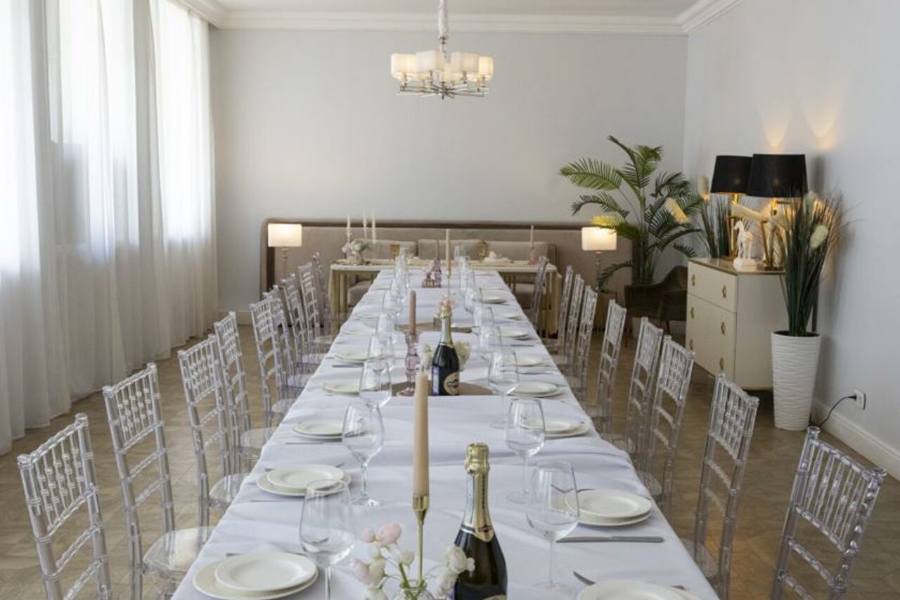 Арендовать Лофт Космос. Зал Президент. Просторное помещение с длинным обеденным столом, покрытым белой скатертью. Стулья прозрачные, на столе расставлены тарелки, бокалы и бутылки. Освещение обеспечивается люстрой и настольными лампами. Комната украшена растениями и декором.