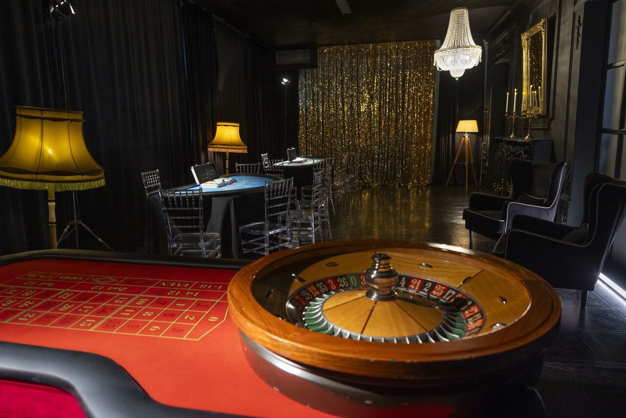 Арендовать Лофт Космос. Зал Президент. Казино в помещении с круглым игровым столом для рулетки на переднем плане. В дальнем углу черный стол с прозрачными стульями и лампами. На заднем плане сверкающая золотая занавеска. Освещение приглушенное, с люстрой и торшером.