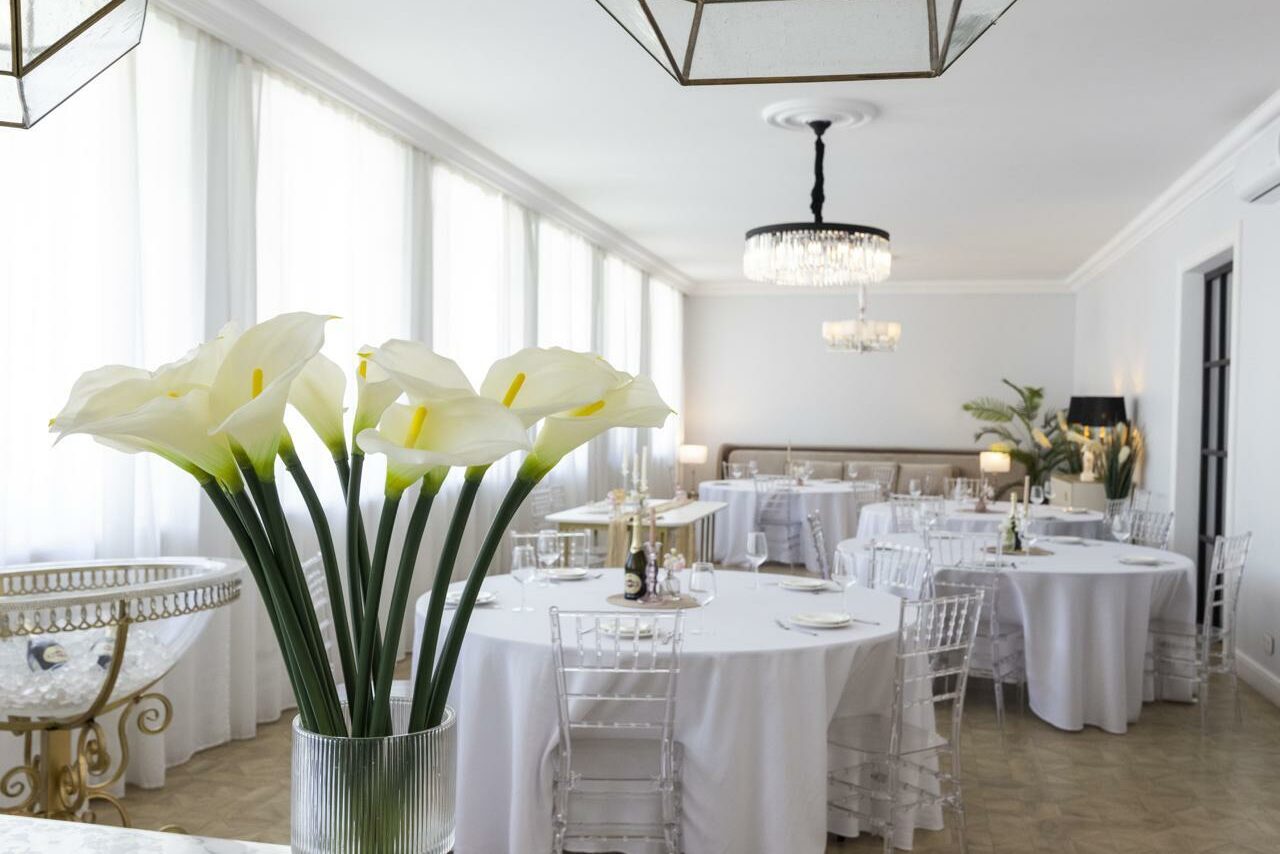 Арендовать Лофт Космос. Зал Президент. Просторный банкетный зал с круглыми столами, накрытыми белыми скатертями, прозрачными стульями, высокими окнами, люстрами и букетами искусственных цветов. Светлые стены, лампы на столах, светлый пол.