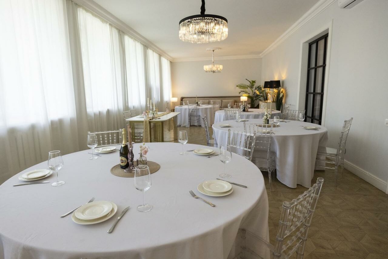 Арендовать Лофт Космос. Зал Президент. Большая, светлая комната с несколькими круглыми столами, накрытыми белыми скатертями. Прозрачные стулья, приборы, бокалы и тарелки на столах. Светильники с кристаллами. Белые стены и большие окна с полупрозрачными шторами. Зеленые растения в углу.