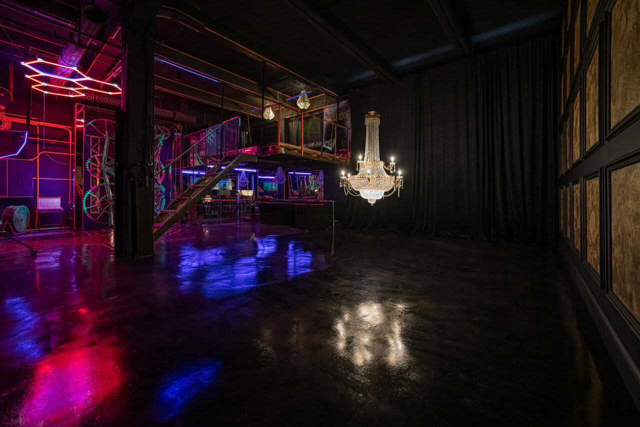 Арендовать Лофт Космос. Зал Ночной Клуб. Частично затемненное помещение с лофтовым интерьером; лестница на второй уровень, стеклянные перила, неоновое освещение, большая люстра, отражающиеся полы, черные стены с занавесками и деревянными панелями.