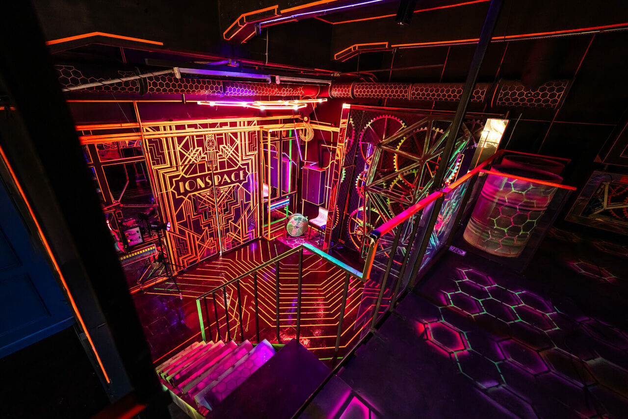 Арендовать Лофт Космос. Зал Ночной Клуб. Лестница с неоновыми акцентами, ведущая вниз. Металлические перила, стены с геометрическим рисунком, светодиодное освещение, создающее футуристическую атмосферу. Яркие неоновые цвета: оранжевый, розовый, зеленый, синий. На стене текст "ON SPACE".