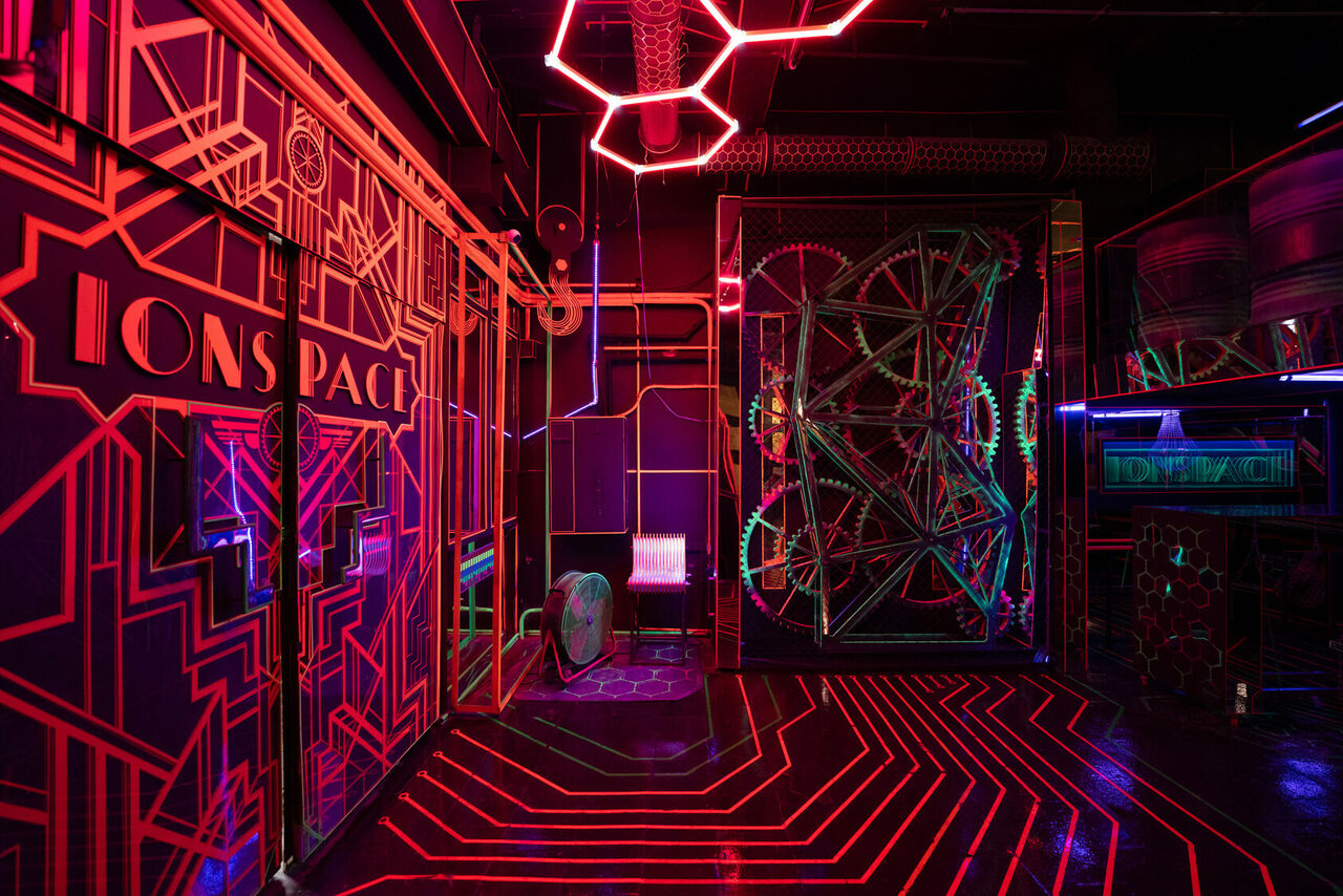 Арендовать Лофт Космос. Зал Ночной Клуб. Футуристическое пространство с геометрической планировкой, неоновая подсветка, металлические конструкции на стенах, неоново-красные и фиолетовые оттенки. Отражающие поверхности, декоративные элементы в виде шестеренок, знак "IONS SPACE".