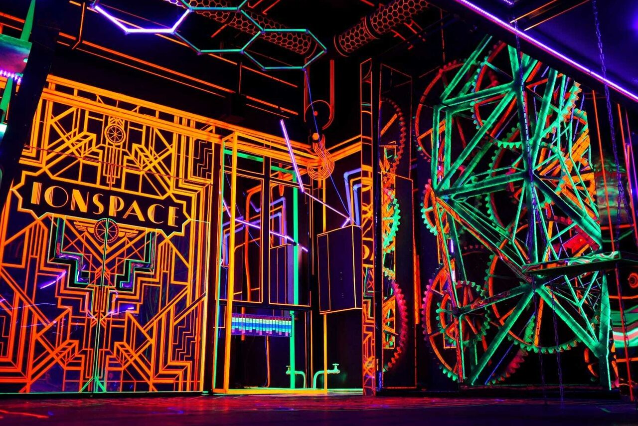 Арендовать Лофт Космос. Зал Ночной Клуб. Интерьер с неоновыми графиками и светящимися линиями на стенах и потолке. Присутствует слово "ONSPACE" на стене. Геометрические узоры и яркие цвета: оранжевый, зеленый, фиолетовый. Элементы декора напоминают шестерни.
