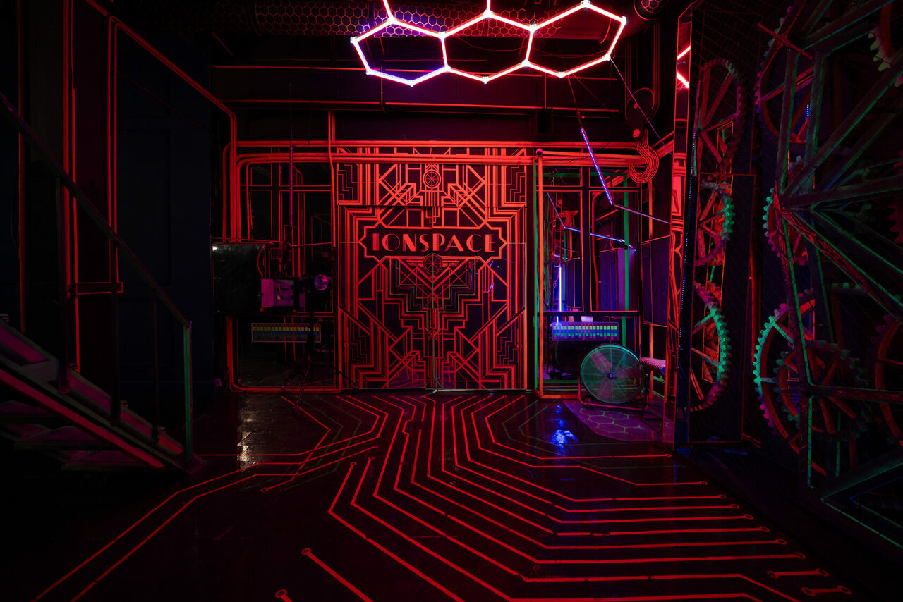 Арендовать Лофт Космос. Зал Ночной Клуб. Темное пространство с неоновыми красными и фиолетовыми линиями на полу и стенах. Металлические конструкции с геометрическими узорами. Светодиодное освещение на потолке в форме шестиугольников. Видны элементы декора в виде шестеренок. Надпись "CONSPACE".