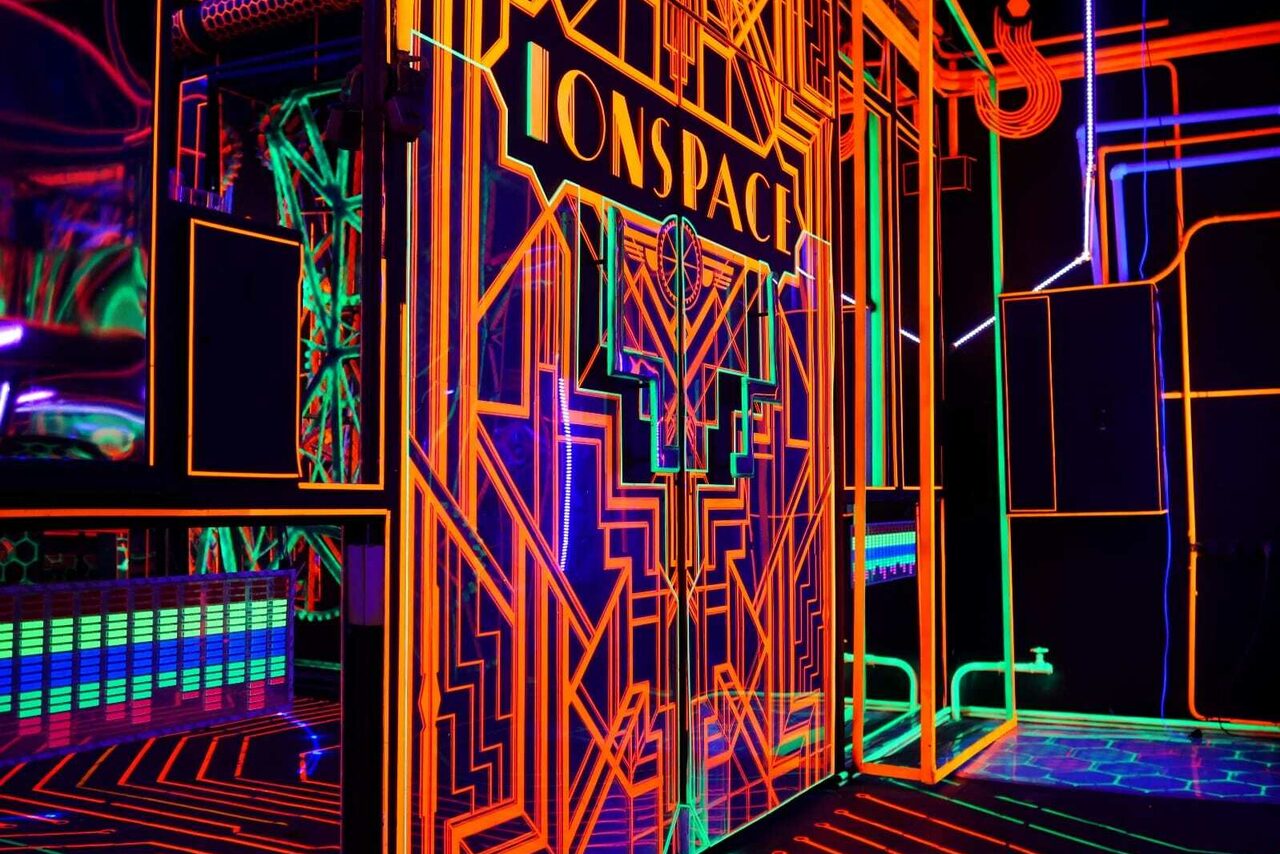 Арендовать Лофт Космос. Зал Ночной Клуб. Пространство с неоновым дизайном в стиле ар-деко. Ярко-оранжевые, зеленые и синие линии на черном фоне. Стены с геометрическим узором, неоновая подсветка. Видна надпись "ONSPACE". Полы синие с яркими линиями.