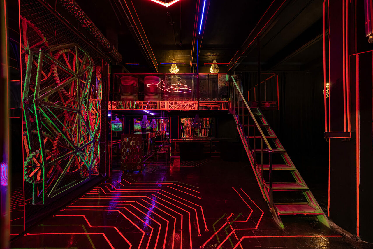 Арендовать Лофт Космос. Зал Ночной Клуб. Темное помещение с неоновым освещением, включая красные и синие световые линии. На первом этаже барная стойка и столики, на втором — металлический балкон с лестницей. Стены декорированы абстрактными узорами, видны геометрические конструкции.