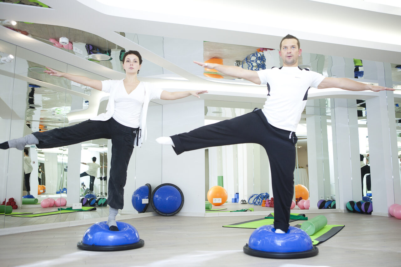 Арендовать Face & Body(Лицо и Тело). Спортивное помещение с зеркальными стенами и светлым деревянным полом. Два человека выполняют упражнения на синих балансировочных полусферах. На полу видны оборудование для фитнеса: гимнастические коврики, мячи и гантели. Яркое накладное освещение.
