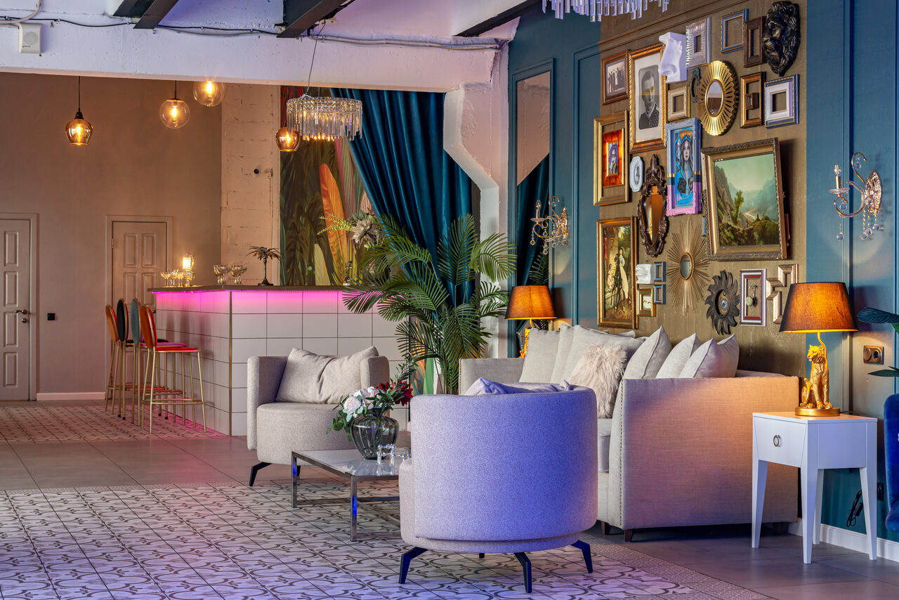 Арендовать Лофт Май. Стильное пространство с верандой. Просторное помещение с зоной отдыха и барной стойкой. Диваны и кресла серых оттенков, журнальный столик, декоративные подушки. Яркие настенные картины и зеркала. Подвесные светильники, лампы. Бар с розовой подсветкой, стулья. Плиточный пол с узором.