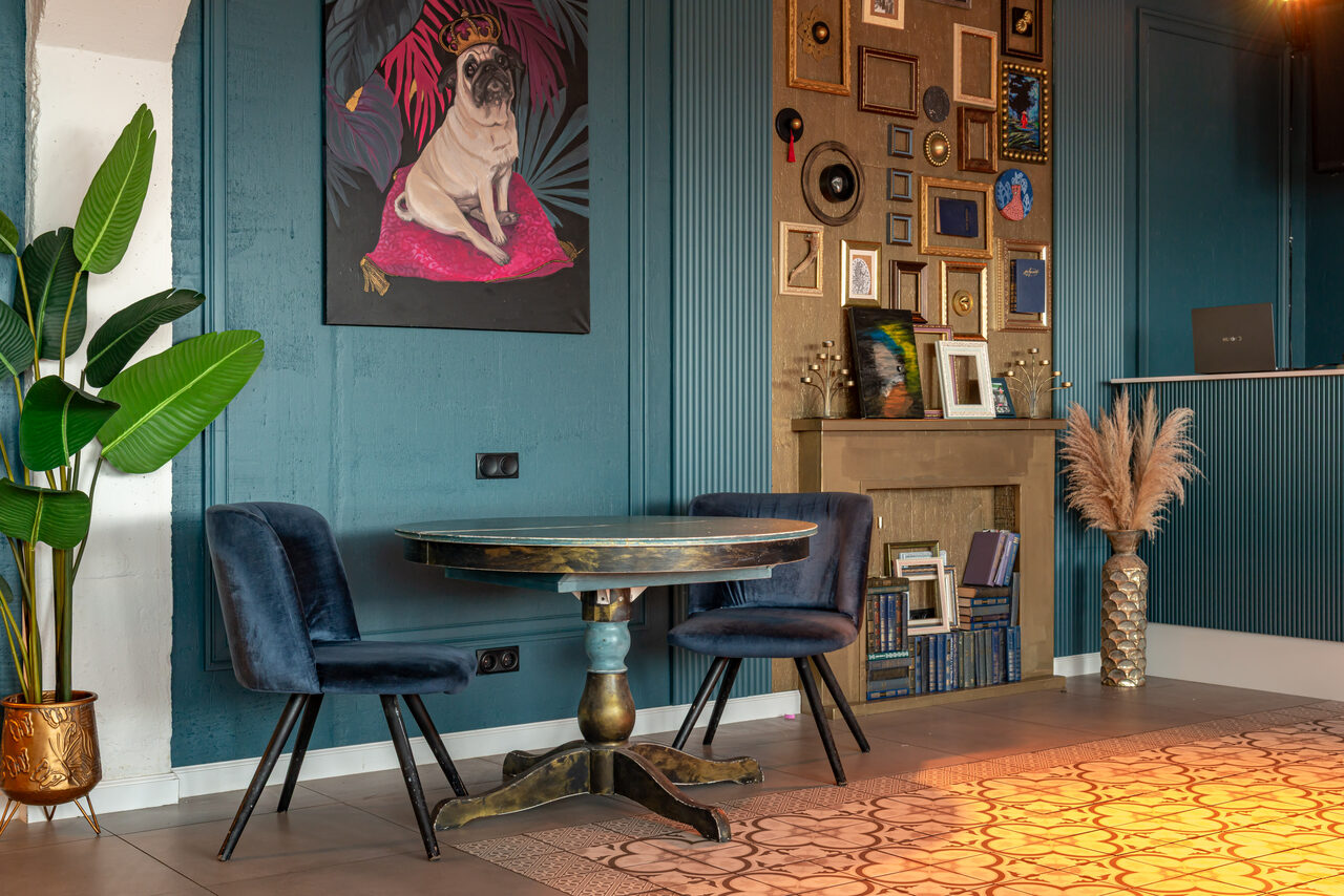 Арендовать Лофт Май. Стильное пространство с верандой. Пространство с синими стенами и декоративными панелями, стол и два синих стула, настенное искусство с изображением собаки, галерея из рамок, растение в горшке, декоративная каминная полка с книгами и аксессуарами, светлый пол с узорчатым ковром.