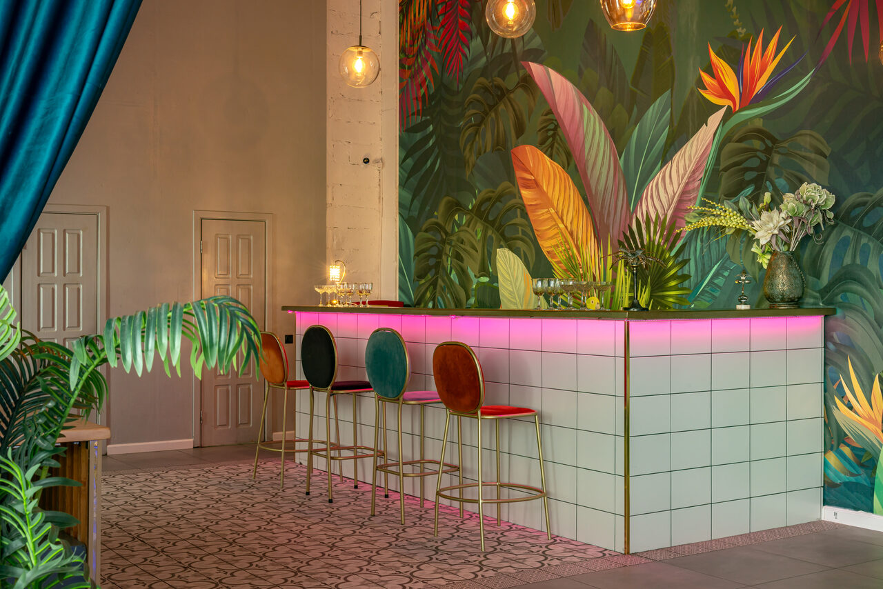 Арендовать Лофт Май. Стильное пространство с верандой. Барная стойка с плиткой светлого цвета, подсветка розовым светом. Четыре высоких стула с цветными сиденьями. Цветочная стеновая роспись. Подвесные светильники. Угловое пространство, декоративные растения. Пол с узором. Два закрытых прохода.