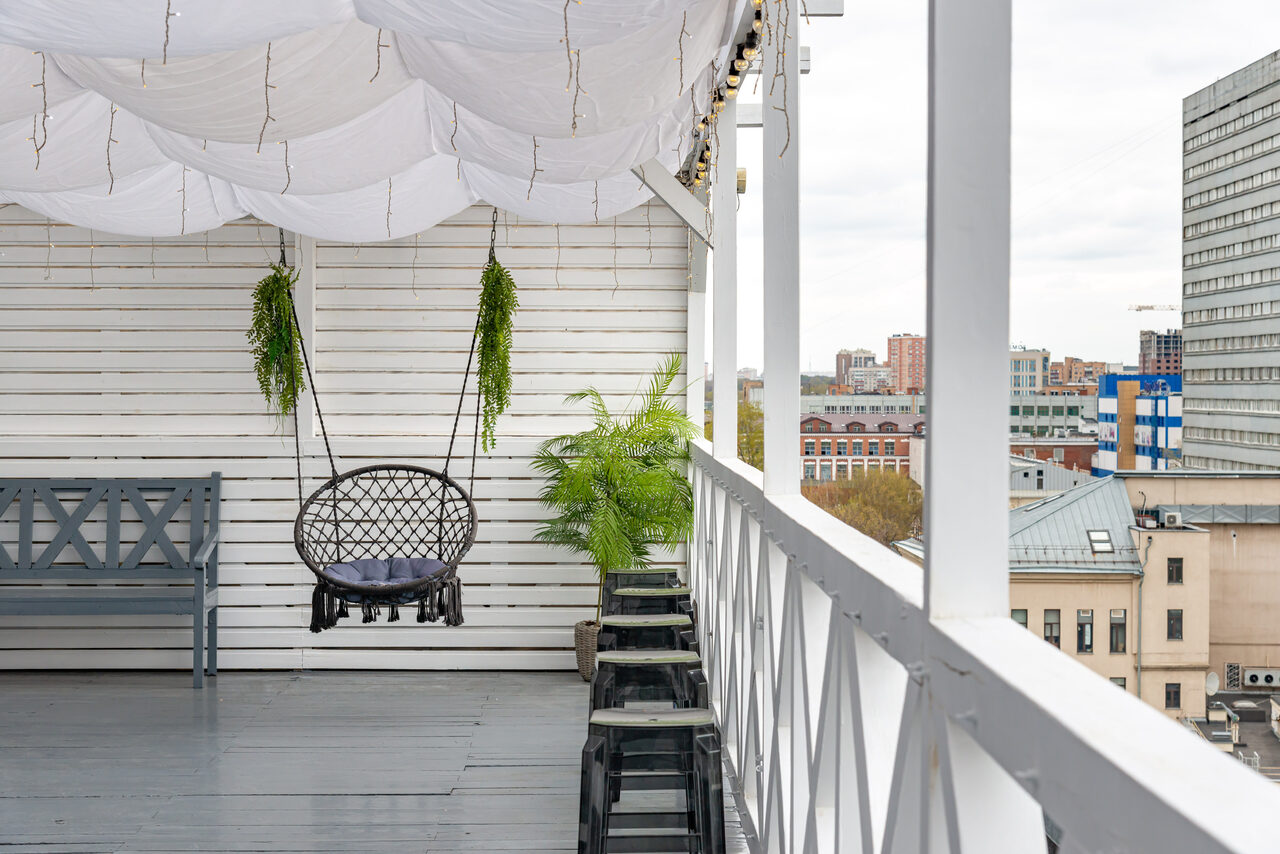 Арендовать Лофт Май. Стильное пространство с верандой. Терраса с деревянным полом и стенами, навесом из белого текстиля. Подвесное кресло и скамейка, вокруг несколько растений в горшках. Декоративные световые гирлянды. Вид на городские здания.