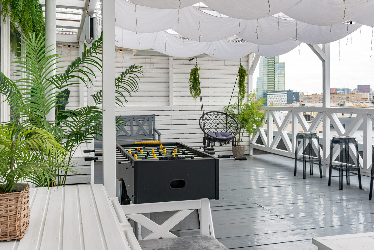 Арендовать Лофт Май. Стильное пространство с верандой. Терраса с белыми деревянными стенами и полом, оснащена настольным футболом, стульями из черного металла и плетеным креслом. Подвешены белые ткани, горшки с зелеными растениями. Панорамный вид на город.