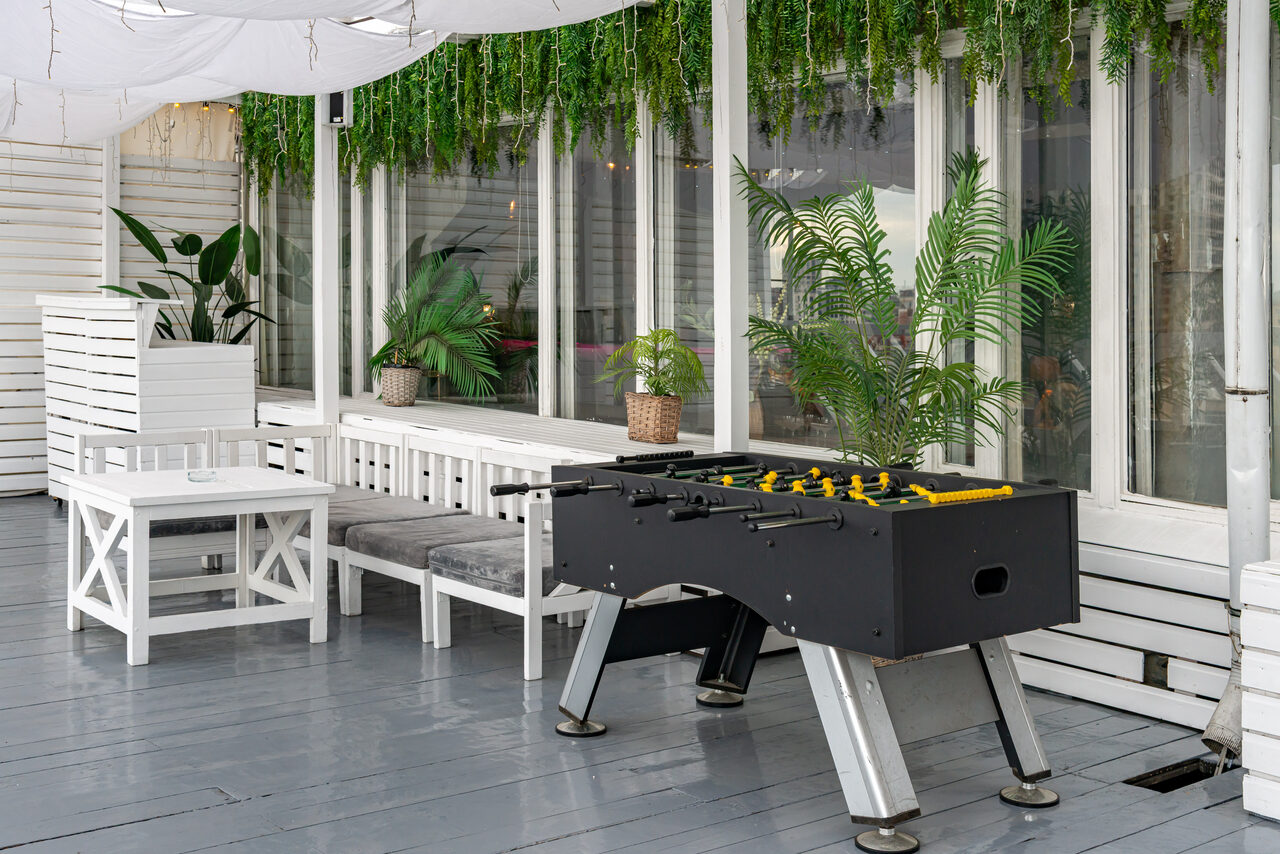 Арендовать Лофт Май. Стильное пространство с верандой. Просторная веранда с деревянным полом серого цвета. По периметру расположены белые деревянные диваны с серыми подушками, белые столы и растения в горшках. Есть настольный футбол. На потолке светлый тент, на стенах декоративная зелень.