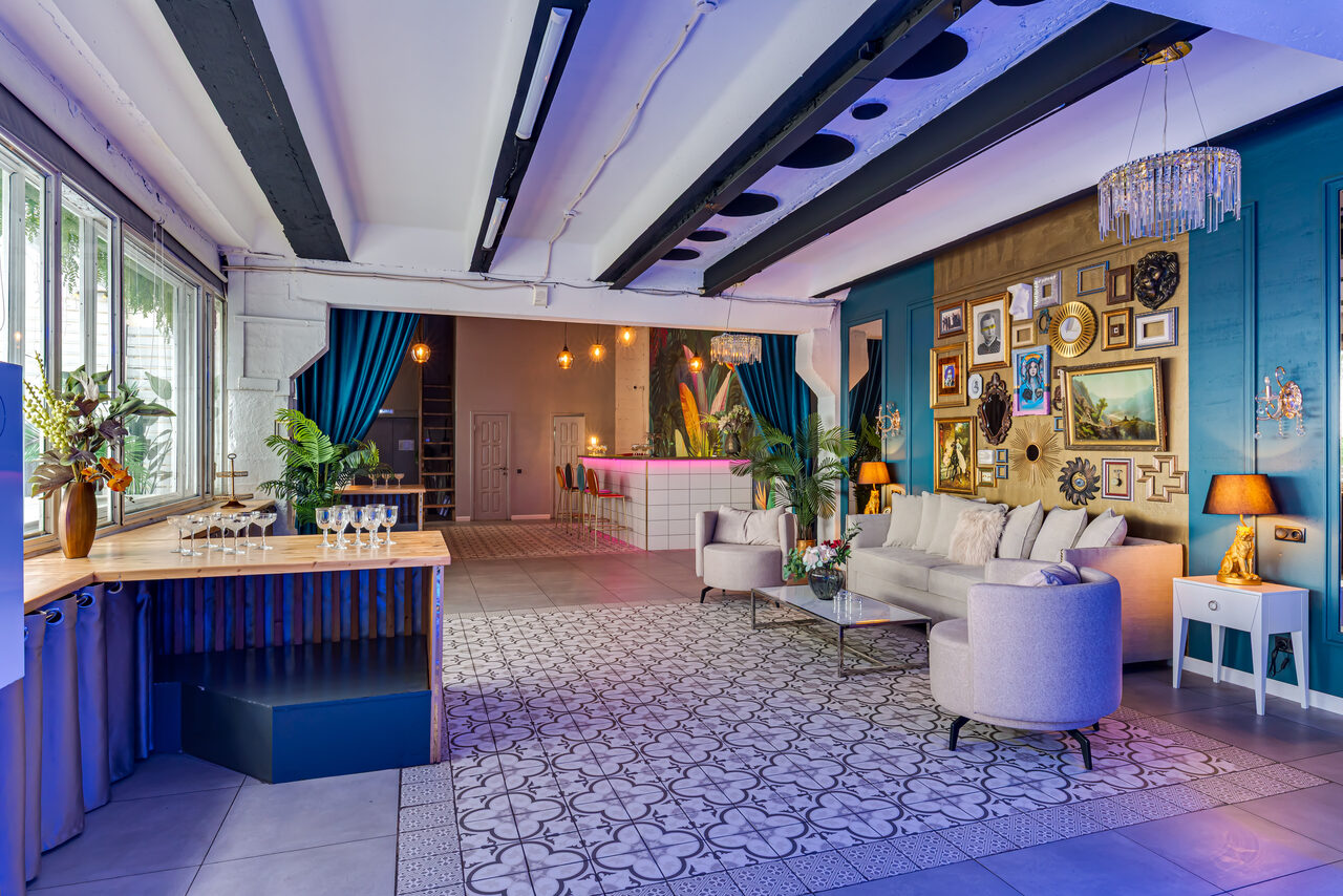 Арендовать Лофт Май. Стильное пространство с верандой. Просторное помещение с зонами для отдыха и бара. Белый диван и кресла на ковре с узором, столик. Барная стойка с бокалами, деревянные и мраморные детали. Стена с рамками и картинами. Потолок с балками, большие окна и плотные шторы. Яркое освещение и люстра.