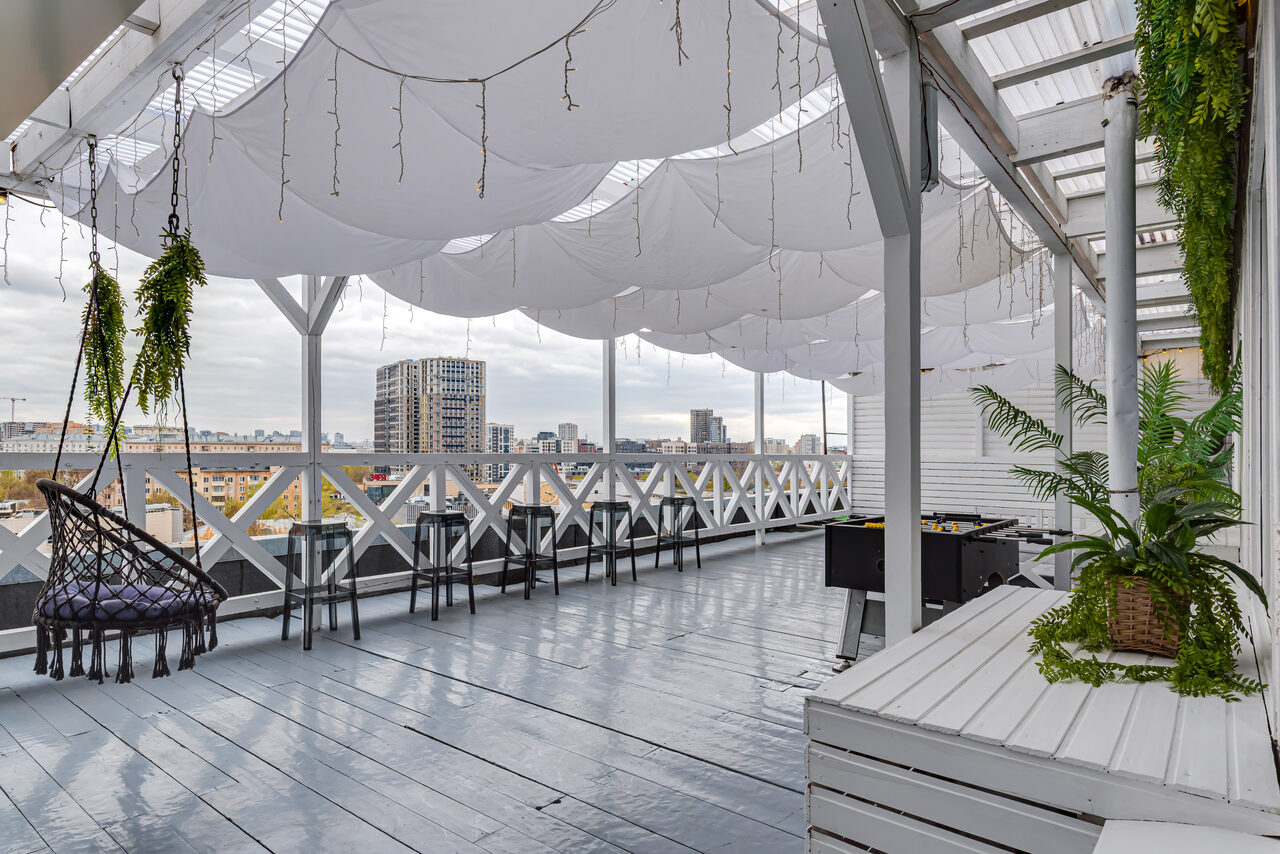 Арендовать Лофт Май. Стильное пространство с верандой. Открытая терраса с деревянным полом и навесом из белой ткани. Вдоль перил стоят высокие барные стулья. В углу расположены качели, стол для настольного футбола и скамейки. На стенах и колоннах висят зелёные растения.