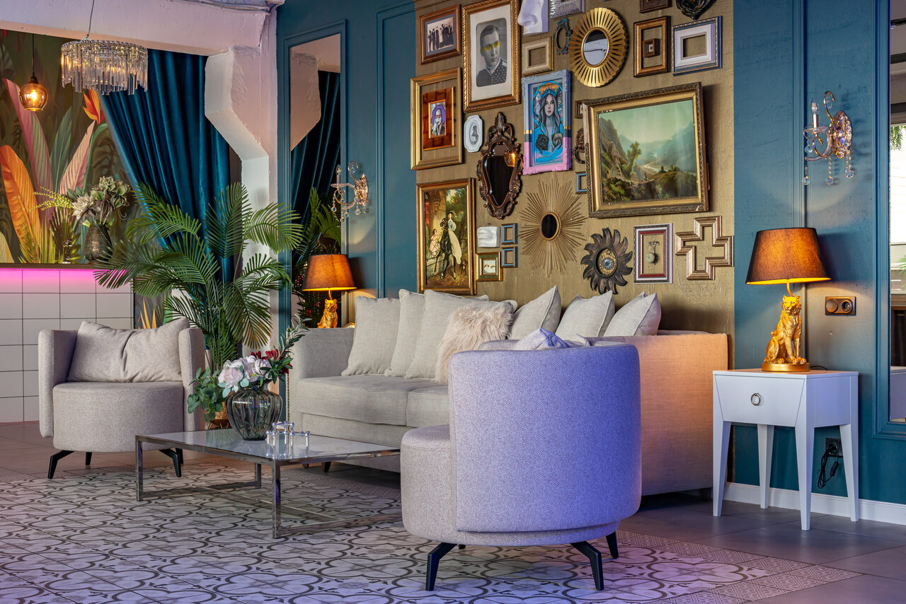 Арендовать Лофт Май. Стильное пространство с верандой. Гостиная с голубыми стенами, украшенными множеством картин в рамках. Мягкий серый диван и кресла, стеклянный столик. Настольные лампы с абажурами, декоративные подушки, растения. Потолочная люстра, плиточный пол с узором, занавес синий.