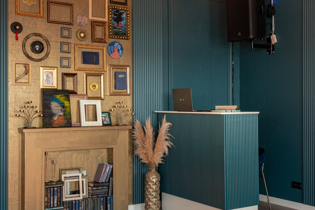 Арендовать Лофт Май. Стильное пространство с верандой. Каморка с отделкой в синих и бежевых тонах, рифленые синие панели, на стене галерея из рамок, столик с книгами и ноутбуком, декоративный камин с книгами в нижней части, ваза с сухими цветами, настенная колонка.