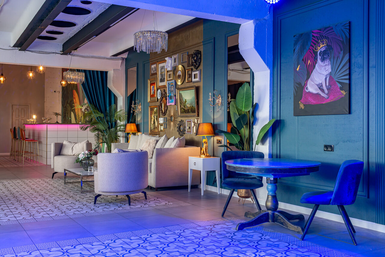 Арендовать Лофт Май. Стильное пространство с верандой. Пространство в лофт-стиле: открытая планировка, зона отдыха с мягкими креслами и столом, барная стойка с подсветкой, декоративные элементы на стенах, картина, разнообразные светильники, синие и белые тона, крупноформатная плитка на полу.