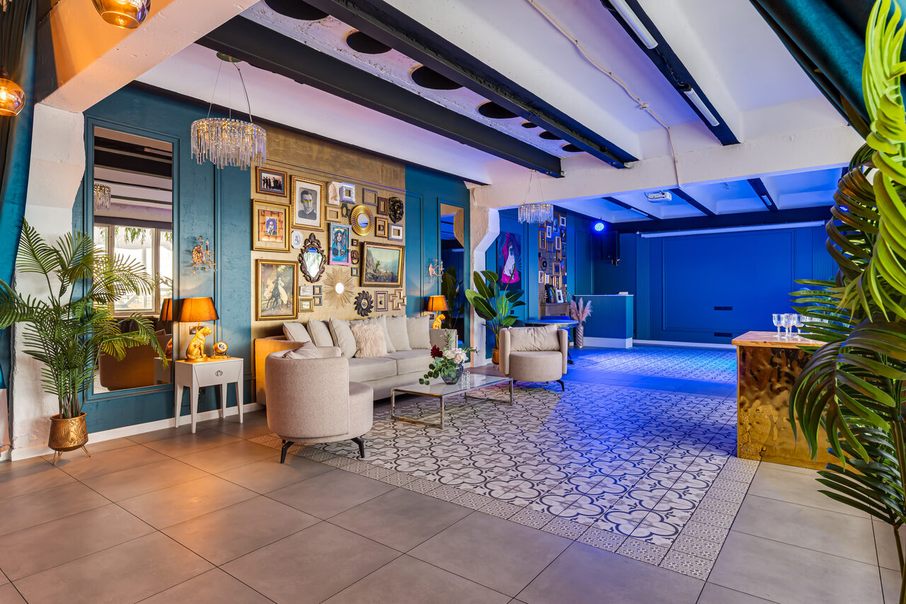 Арендовать Лофт Май. Стильное пространство с верандой. Просторное помещение с мебелью для отдыха: диван и кресла кремового цвета, журнальный столик. Стены украшены картинами, голубые и золотистые акценты. Плиточный пол с узором, зеркала, комнатные растения, декоративное освещение.