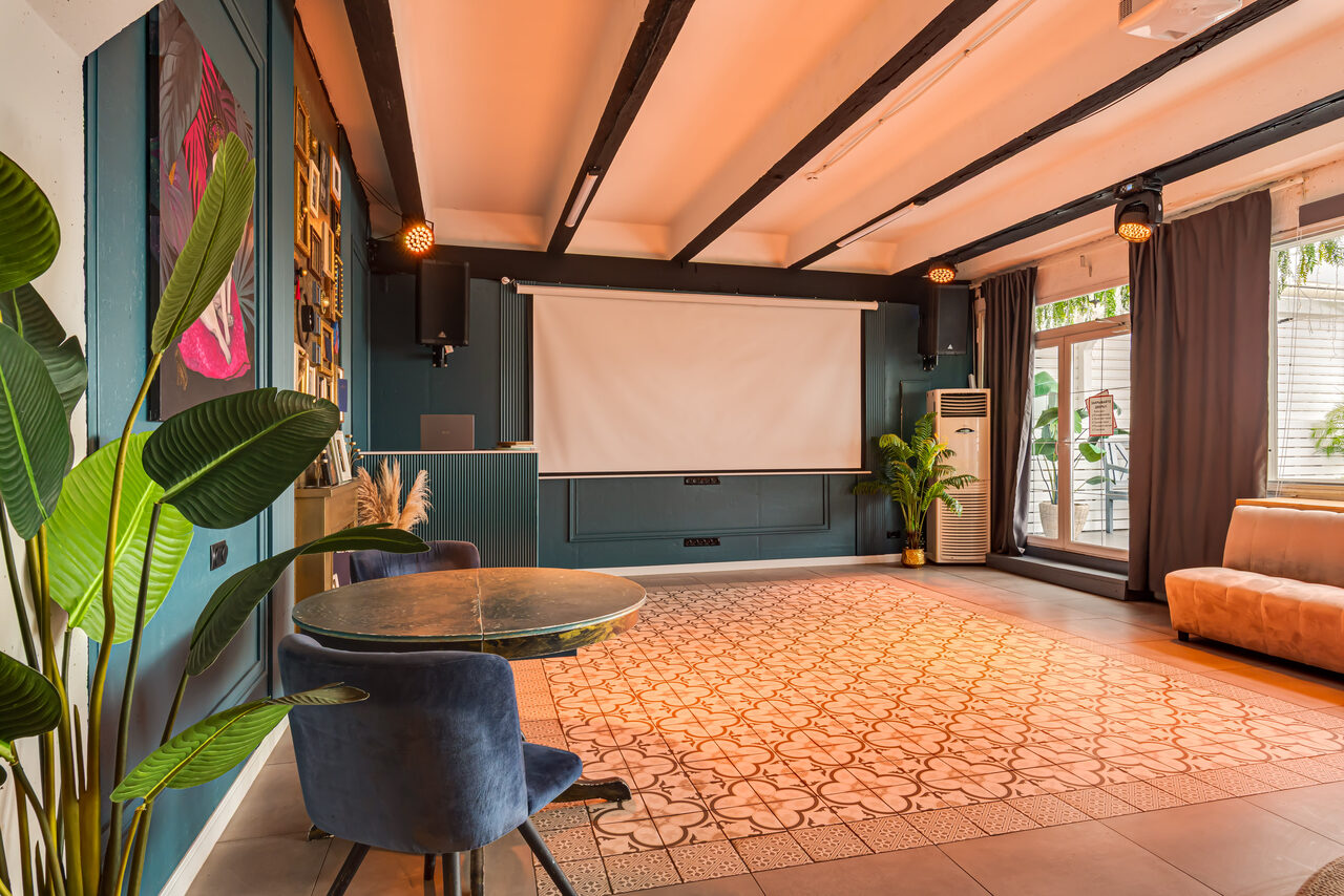 Арендовать Лофт Май. Стильное пространство с верандой. Просторное помещение с проектором и экраном на стене. Потолок с чёрными балками, пол с узорчатой плиткой. В помещении стол с синим креслом, диван, растения, окна с тёмными шторами. Освещение с потолочными светильниками.