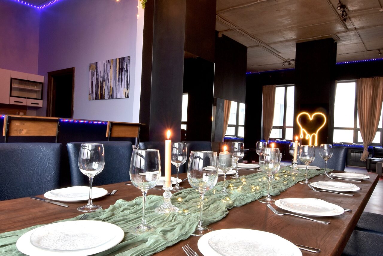 Арендовать Элегантный лофт с хорошим звуком и балконом. Просторное помещение с деревянным столом, сервированным бокалами и тарелками. На стене картина, рядом открытая кухня с мебелью. Потолок с накладным освещением, стены светлые. Светодиодная подсветка по периметру, большое окно со шторами.